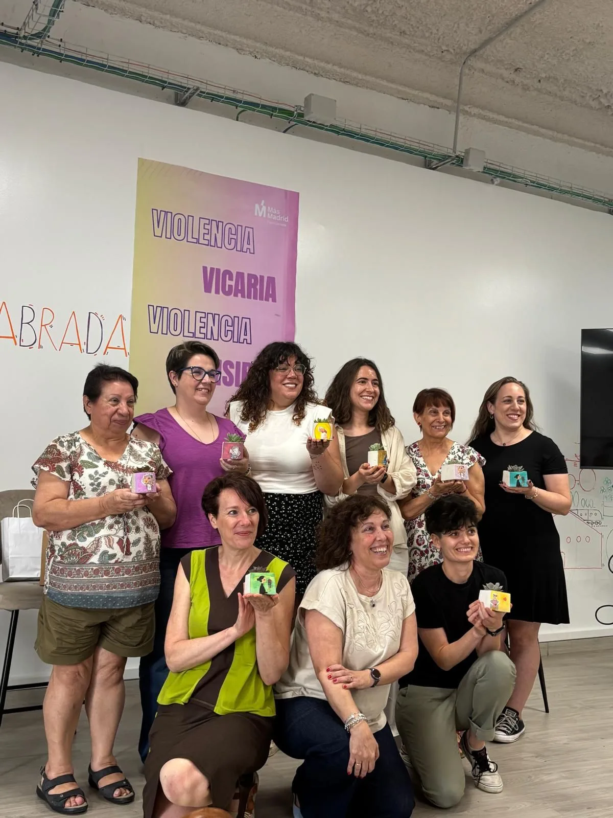 Mujeres posando con pequeñas macetas ante un cartel sobre violencia vicaria en el encuentro de microteatro